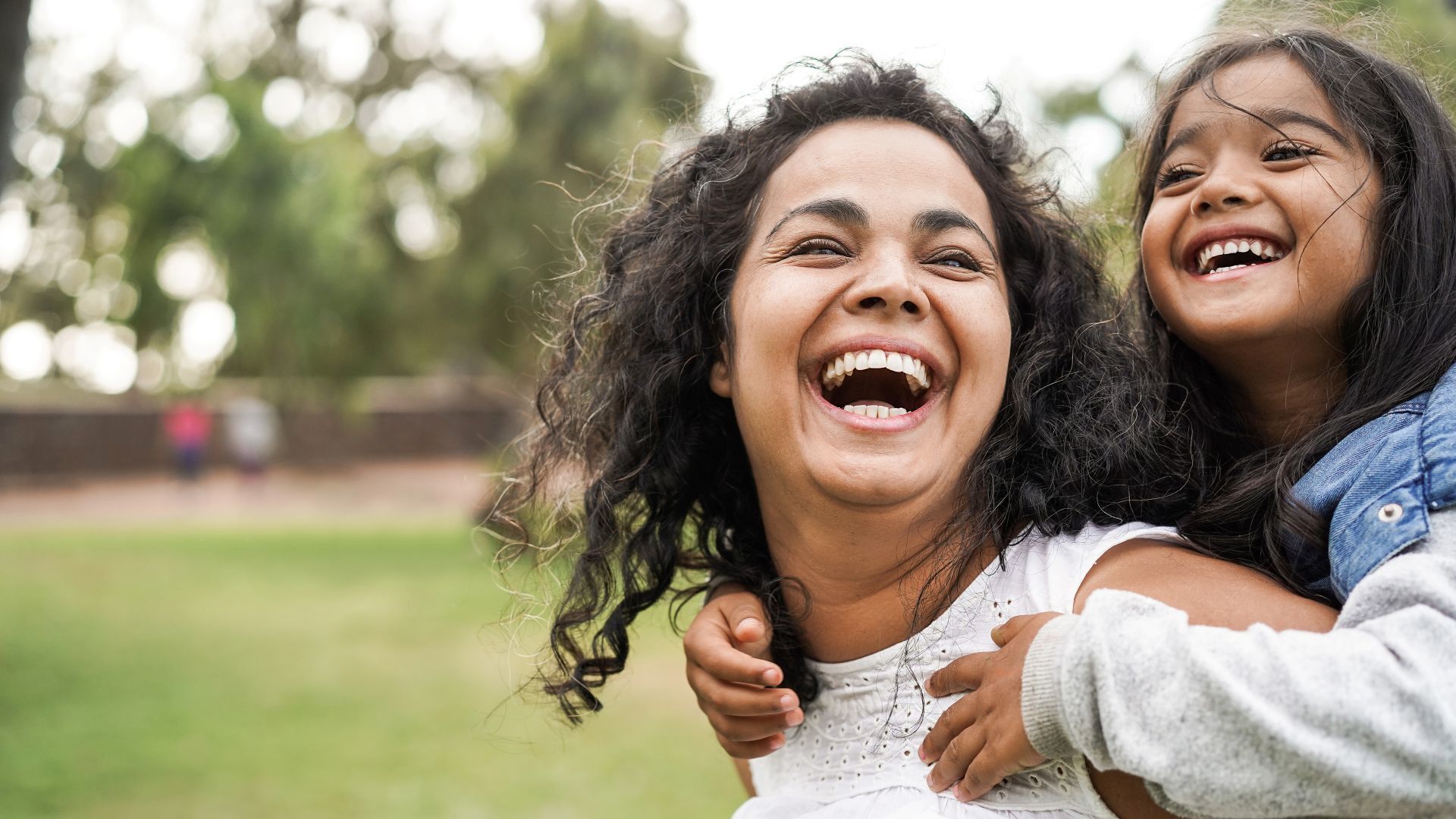 A woman with curly hair smiles while carrying a child on their back, set against a green park background.