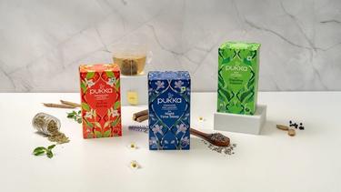 Three Pukka tea boxes are displayed on a white surface: red for Active Energy, blue for Night Time Sleep, and green for Digestive Support, with matching herbs and a glass of tea in the background.