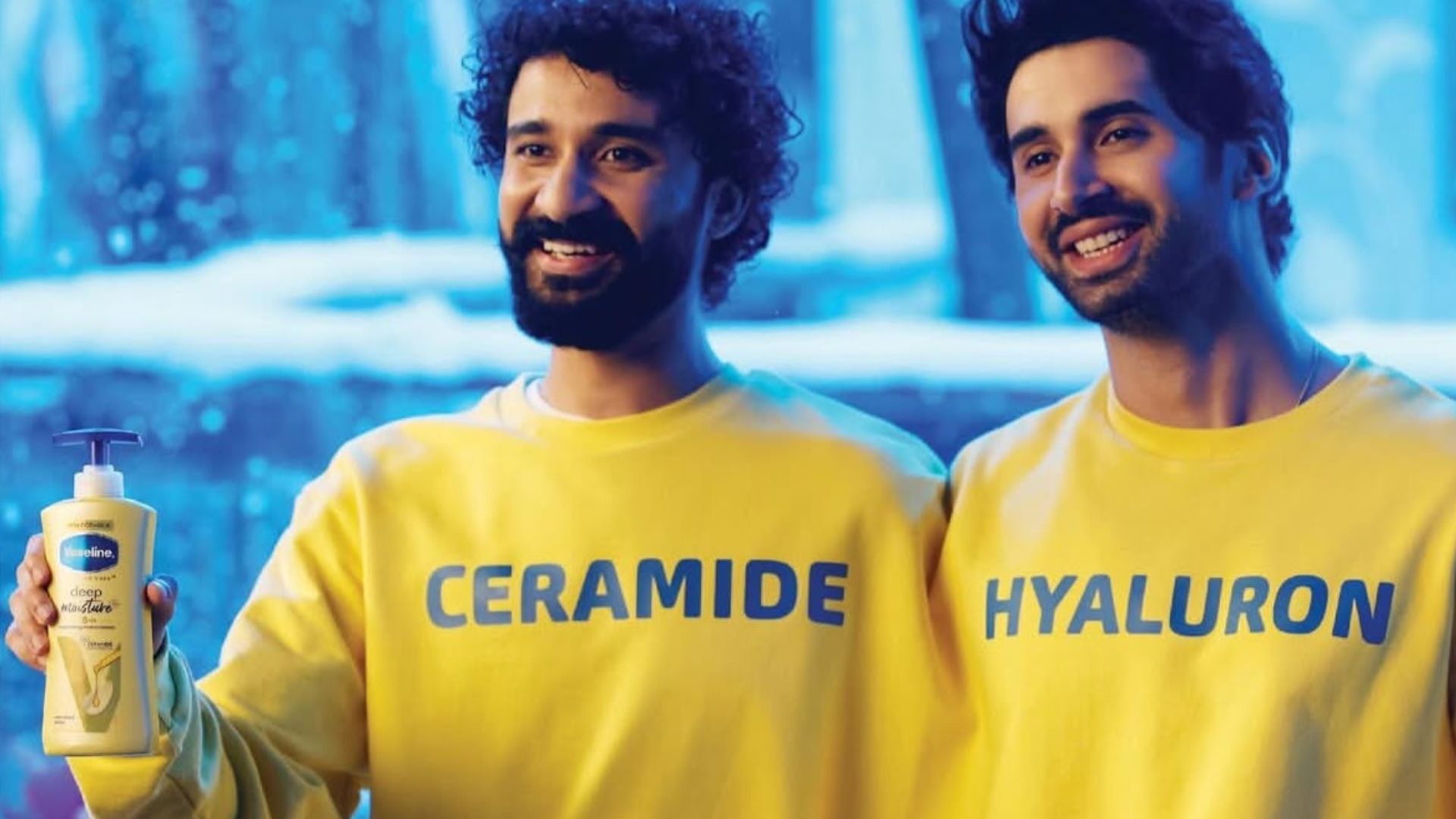 Two smiling men in yellow branded sweatshirts hold a Vaseline Deep Moisture lotion bottle against a blue background
