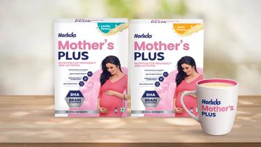 Two packs of Horlicks Mother's Plus nutrition drink in vanilla and kesar flavours featuring a pregnant woman in a pink dress, along with a filled mug displayed on a wooden surface.