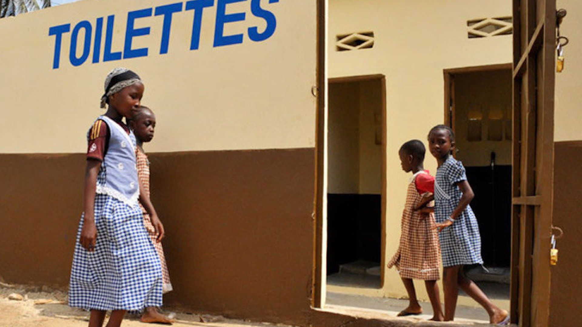 Four young African girls in school uniforms walking into school toilets marked with a painted sign saying ‘toilettes’.