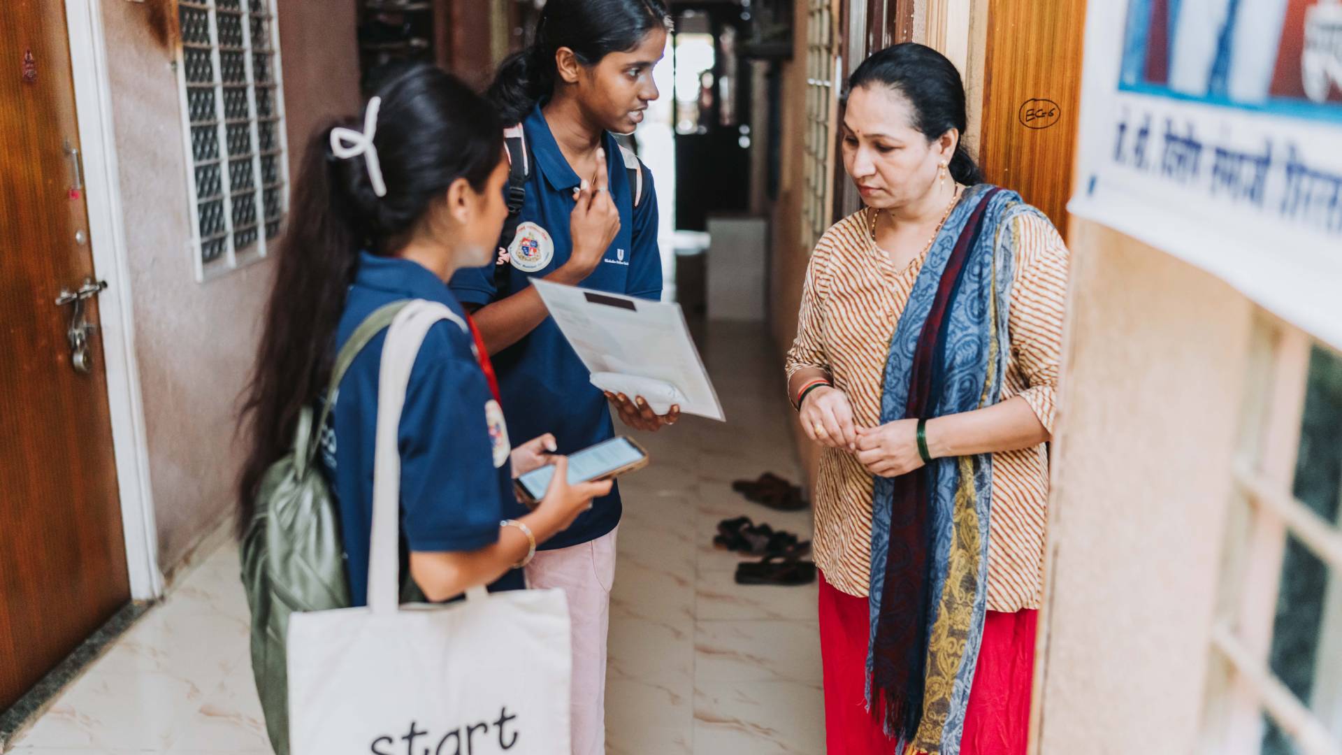 Two Indian students in blue shirts present information on a tablet and clipboard to a woman in a striped shirt with a blue scarf, likely for a survey or community outreach program.