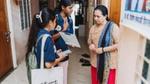 Two Indian students in blue shirts present information on a tablet and clipboard to a woman in a striped shirt with a blue scarf, likely for a survey or community outreach program.