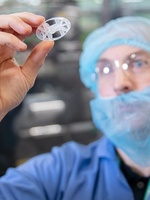 A gloved hand holding a small, circular plastic component in a cleanroom environment with machinery in the background.