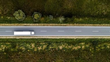 Aerial view of a truck driving along a straight, empty road surrounded by trees on either side.