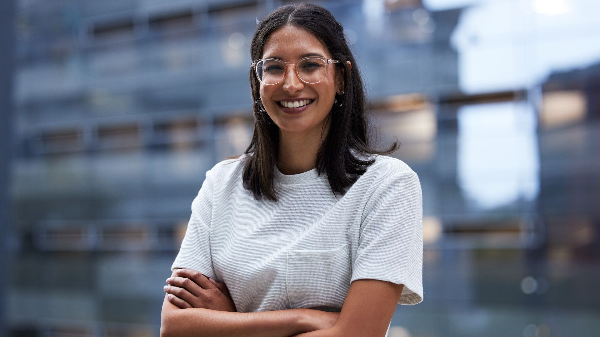 A confident woman in white shirt wearing glasses with her hands crossed smilling to the camera
