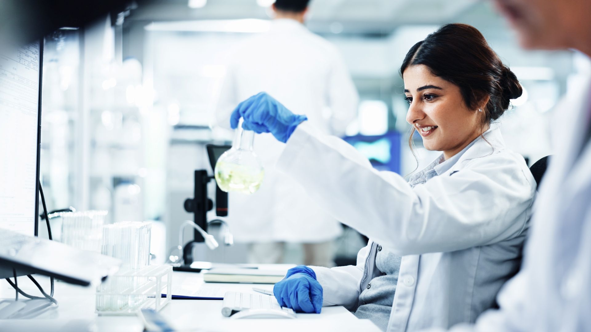 A lab technician examines a solution in a glass flask while working at a laboratory bench with scientific equipment nearby.