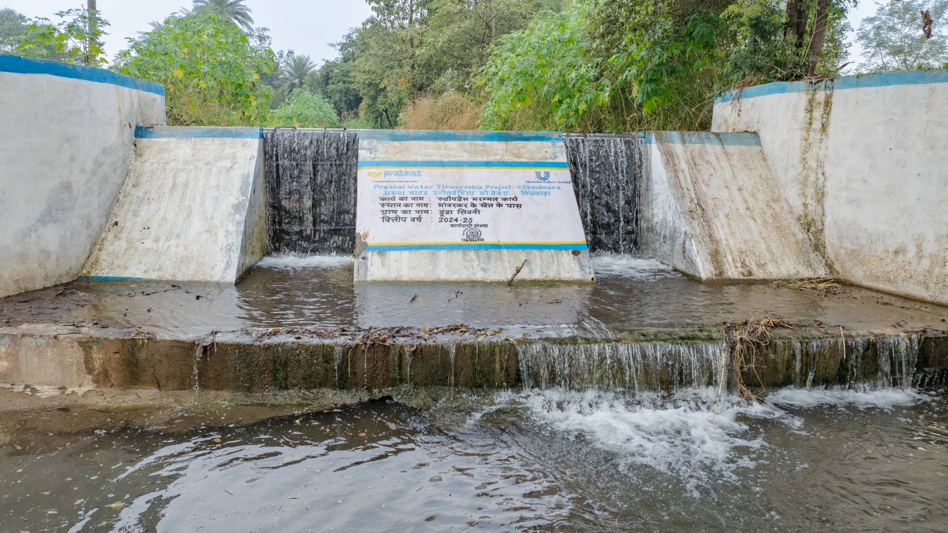 Small dam with signage and water flowing over it amidst greenery.