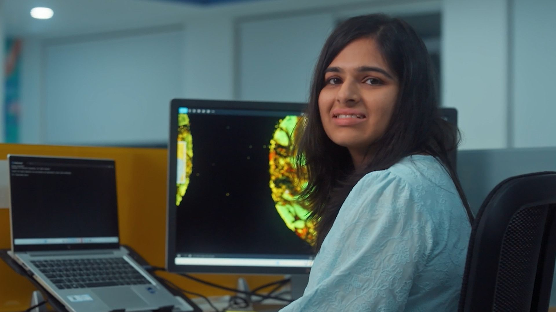 Young professional at a desk smiles toward the camera with data visualizations on her computer screen.