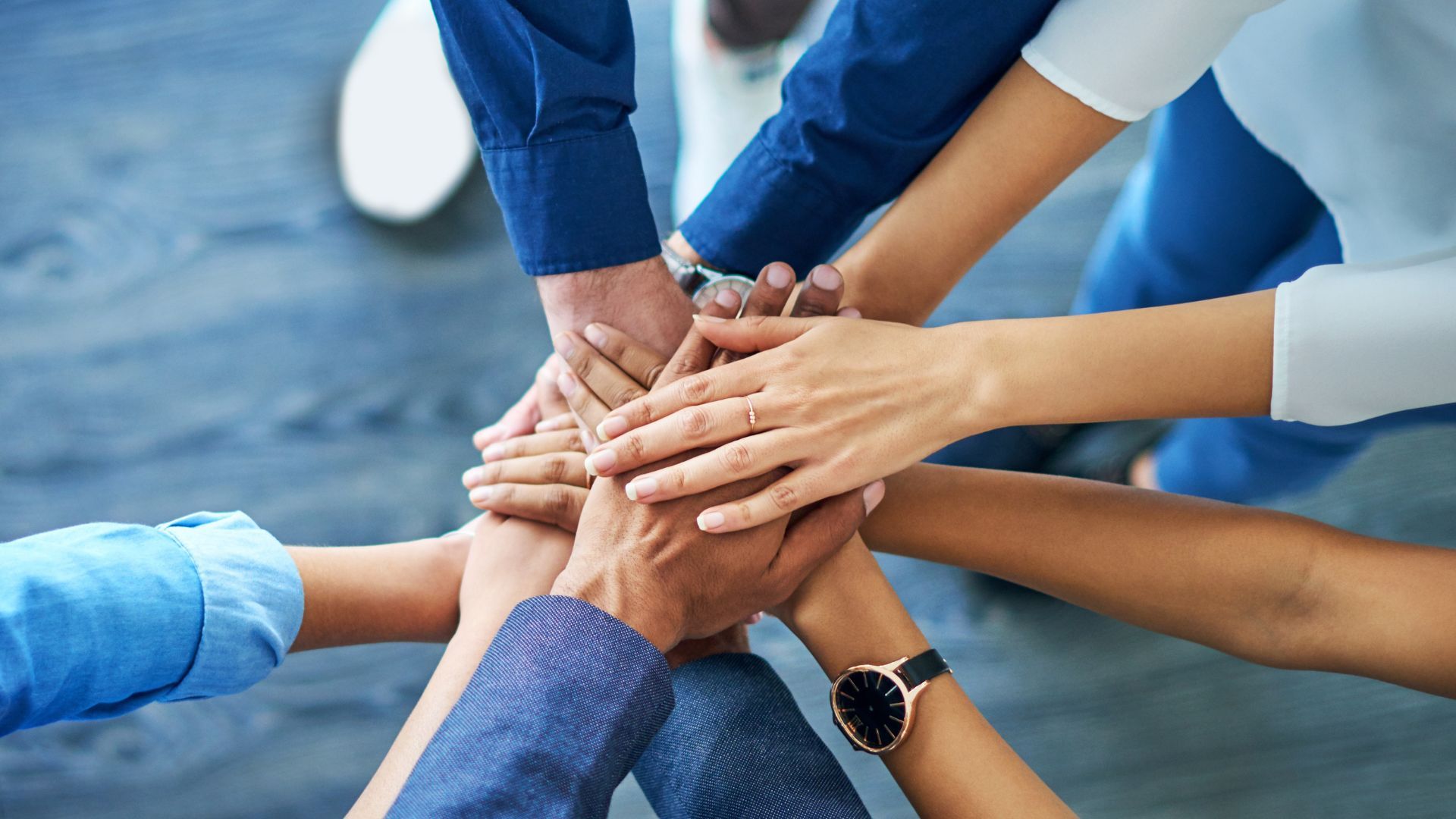  A group of people placing their hands in the centre of a circle in a pile. All are wearing clothes in shades of blue.