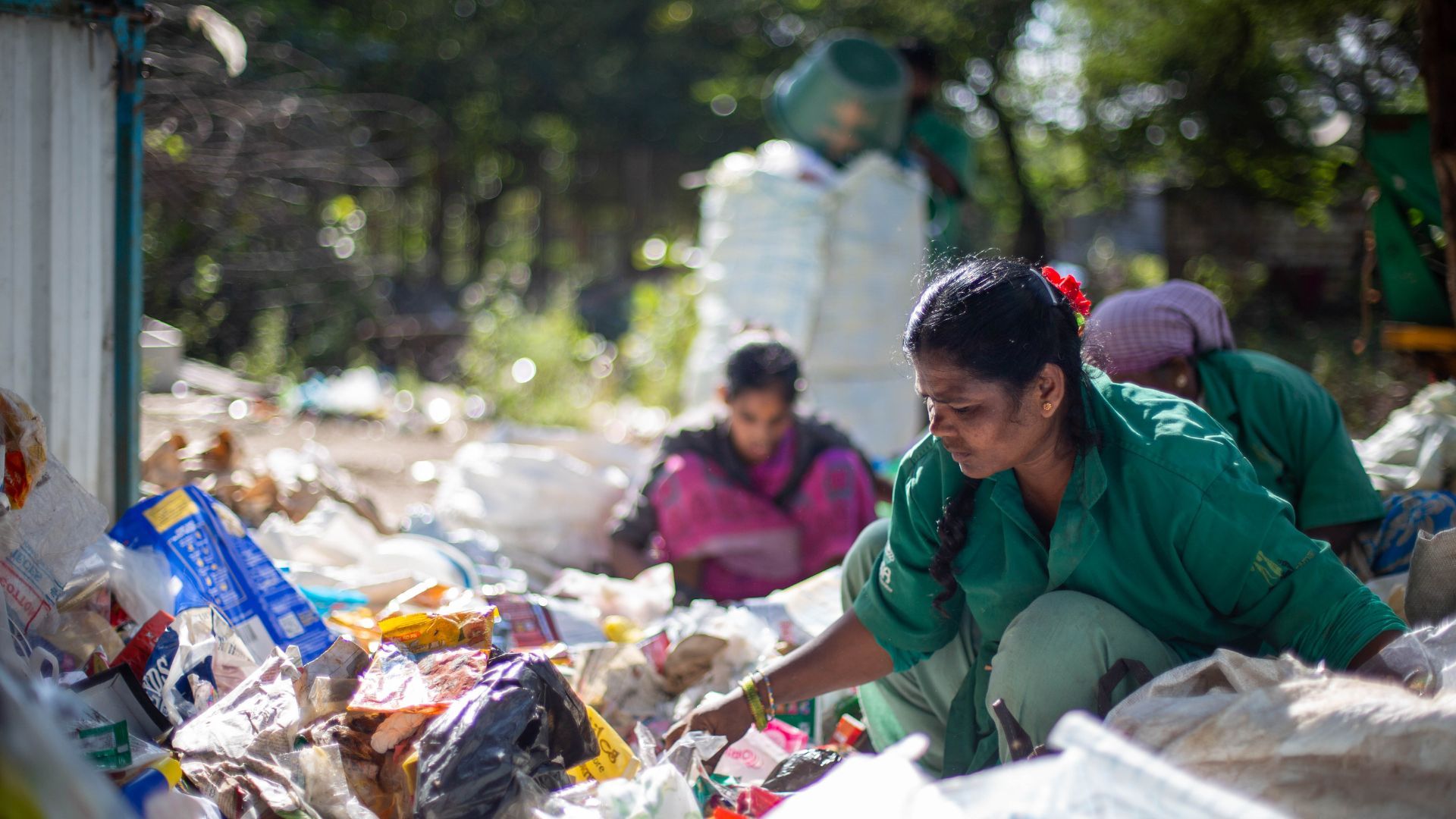 Women in green uniforms sort recyclable waste outdoors at a recycling facility.