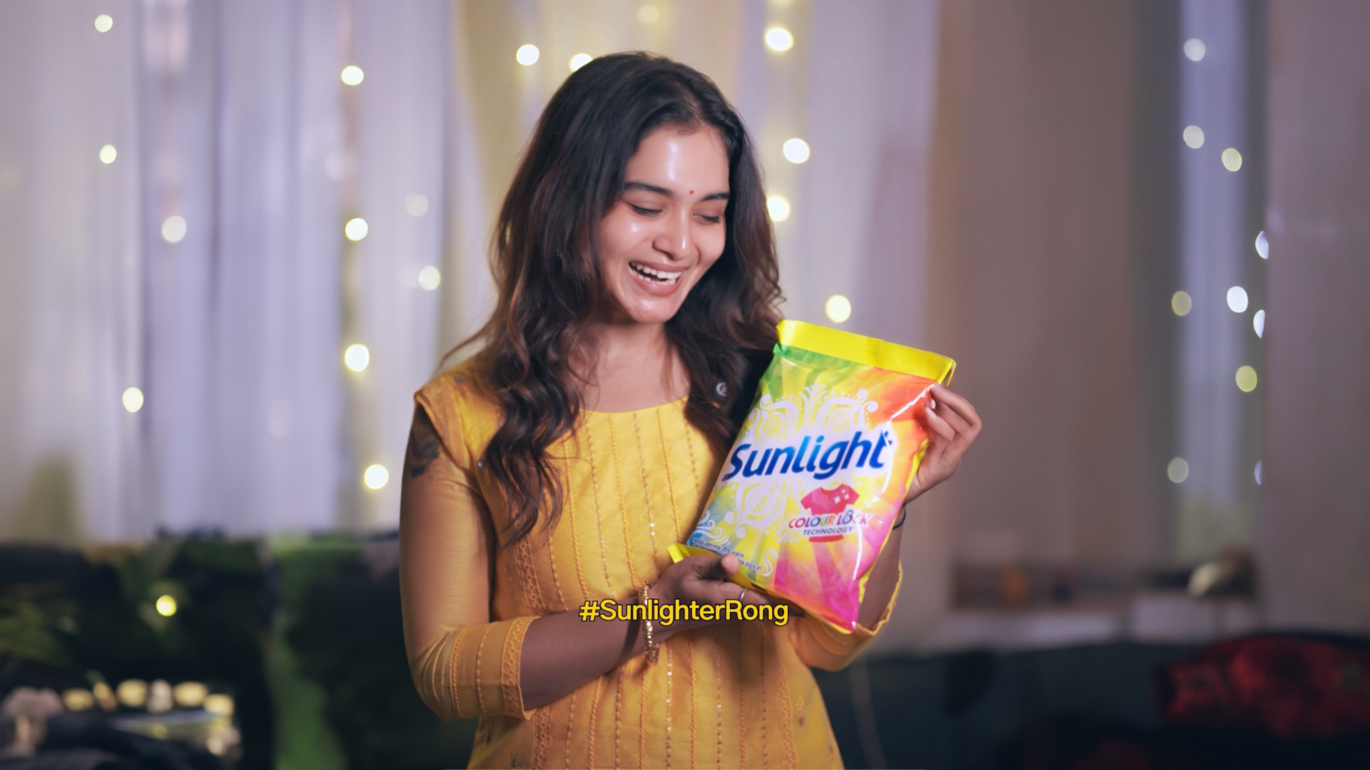 A smiling woman in a yellow outfit holds a colorful packet of Sunlight detergent, with fairy lights in the background.
