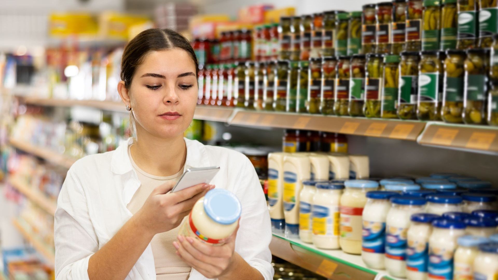 A woman looking at a label on a jar of mayonnaise, standing next to a grocery aisle filled with various products on the shelves.