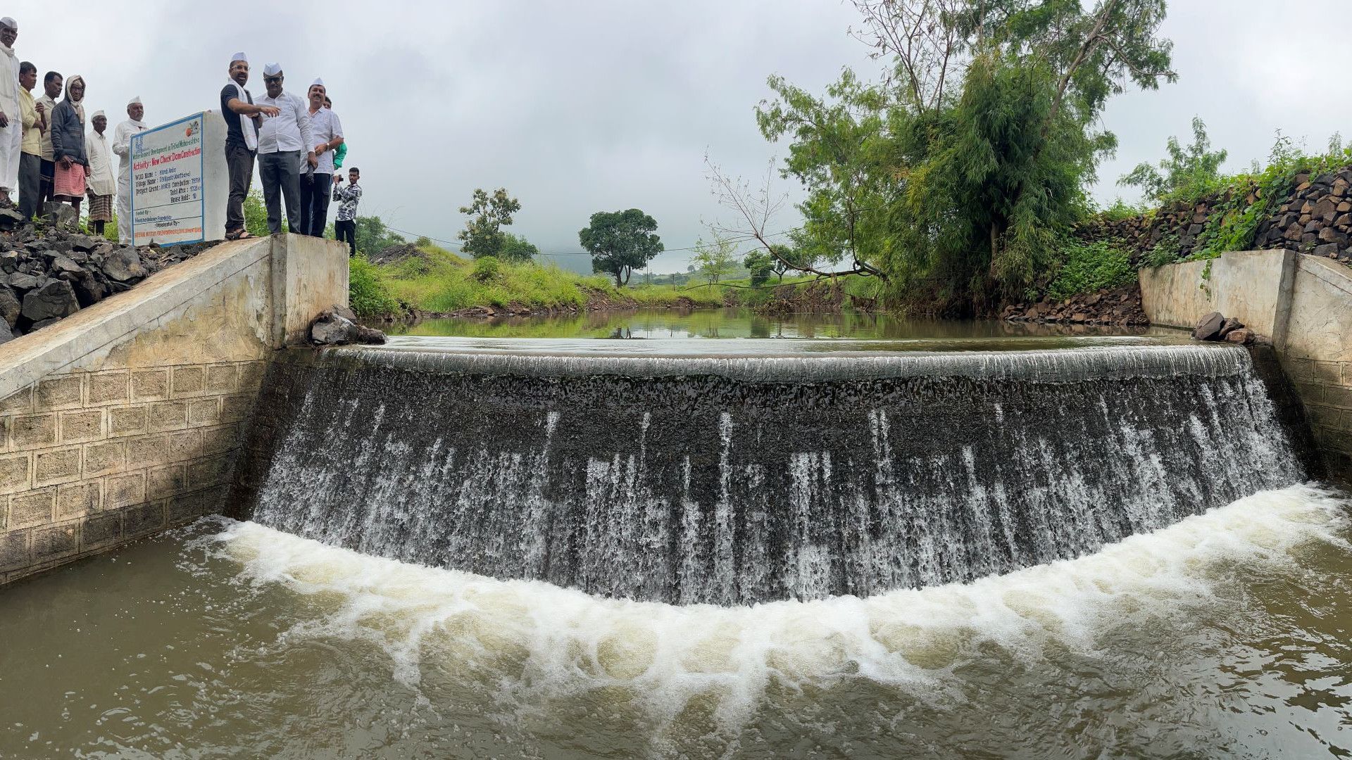 Community members inspect a small concrete check dam with water flowing over the edge.