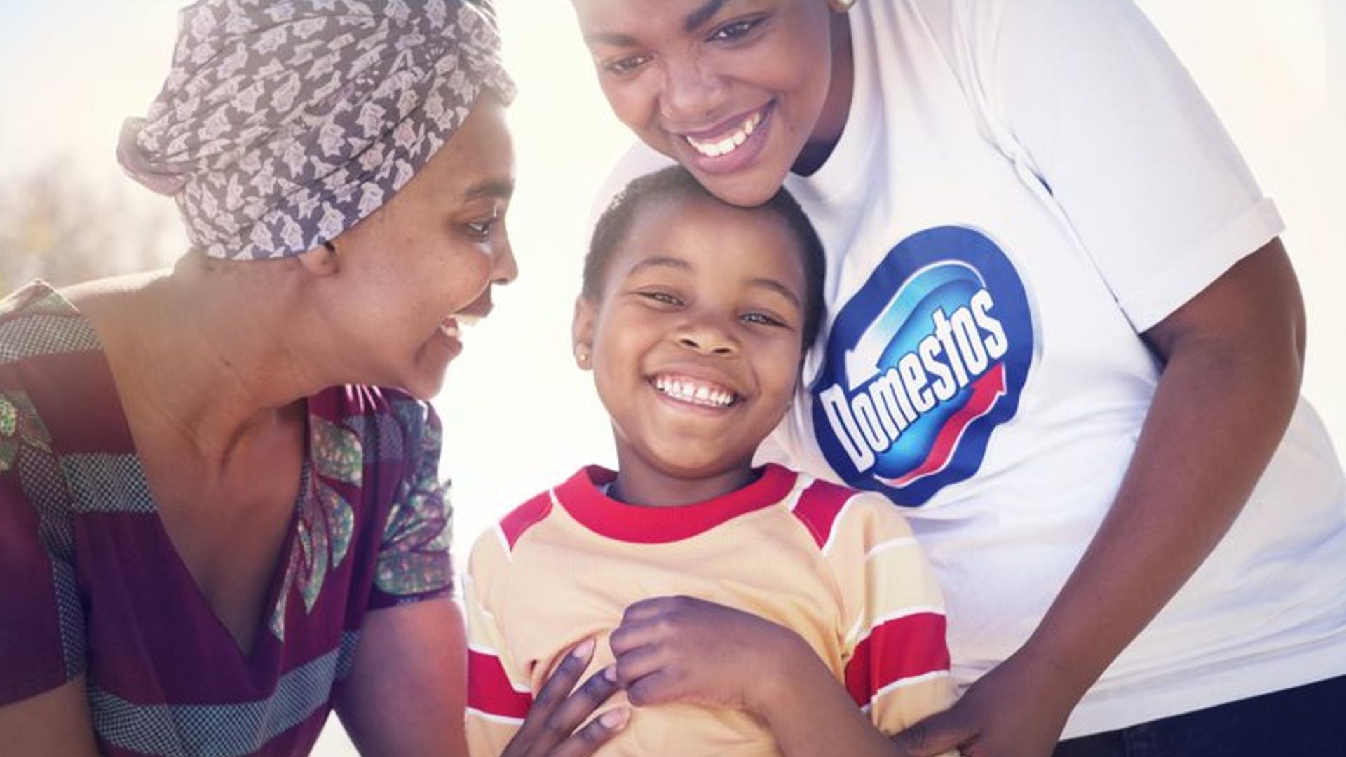 Two women, one wearing a Domestos t-shirt, laughing with a young child