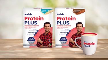 Two packs of Horlicks Protein Plus packs in vanilla and chocolate flavours featuring a man in a red shirt, along with a filled mug displayed on a wooden surface.