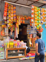 A small street-side shop displaying hanging packets of snacks and jars of candies, with a person standing in front of the shop.