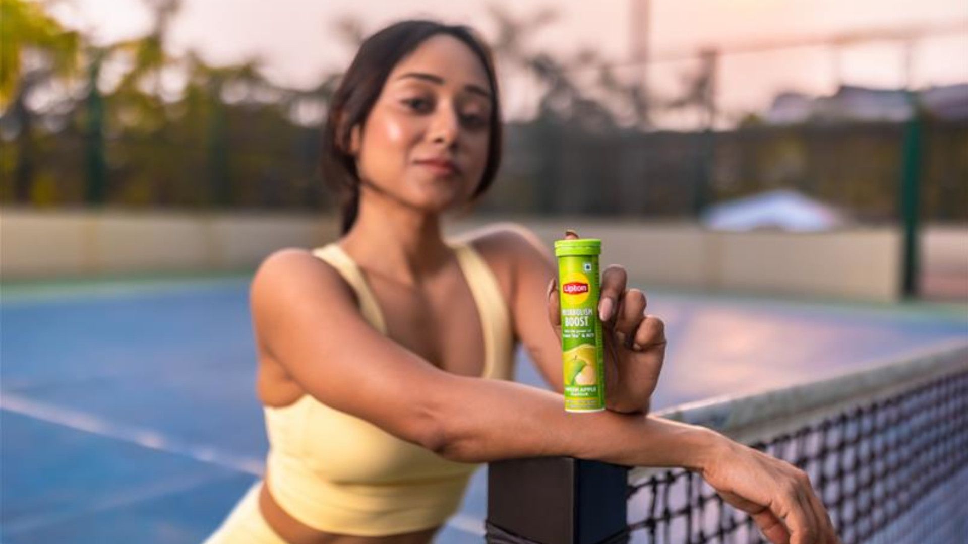 A woman in a yellow sports outfit stands on a tennis court at sunset, leaning on a net post. She's holding a green Lipton Energy Boost bottle, smiling slightly.