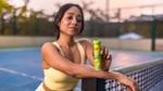 A woman in a yellow sports outfit stands on a tennis court at sunset, leaning on a net post. She's holding a green Lipton Energy Boost bottle, smiling slightly.