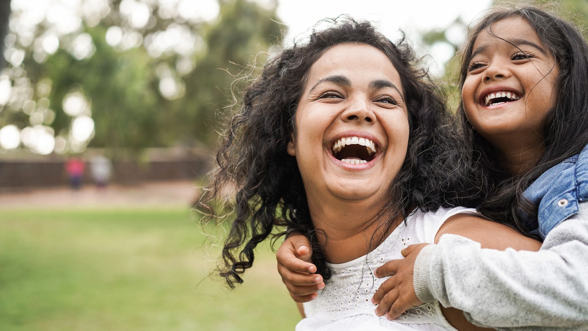A mother is giving her daughter a piggy back and both are laughing in the park