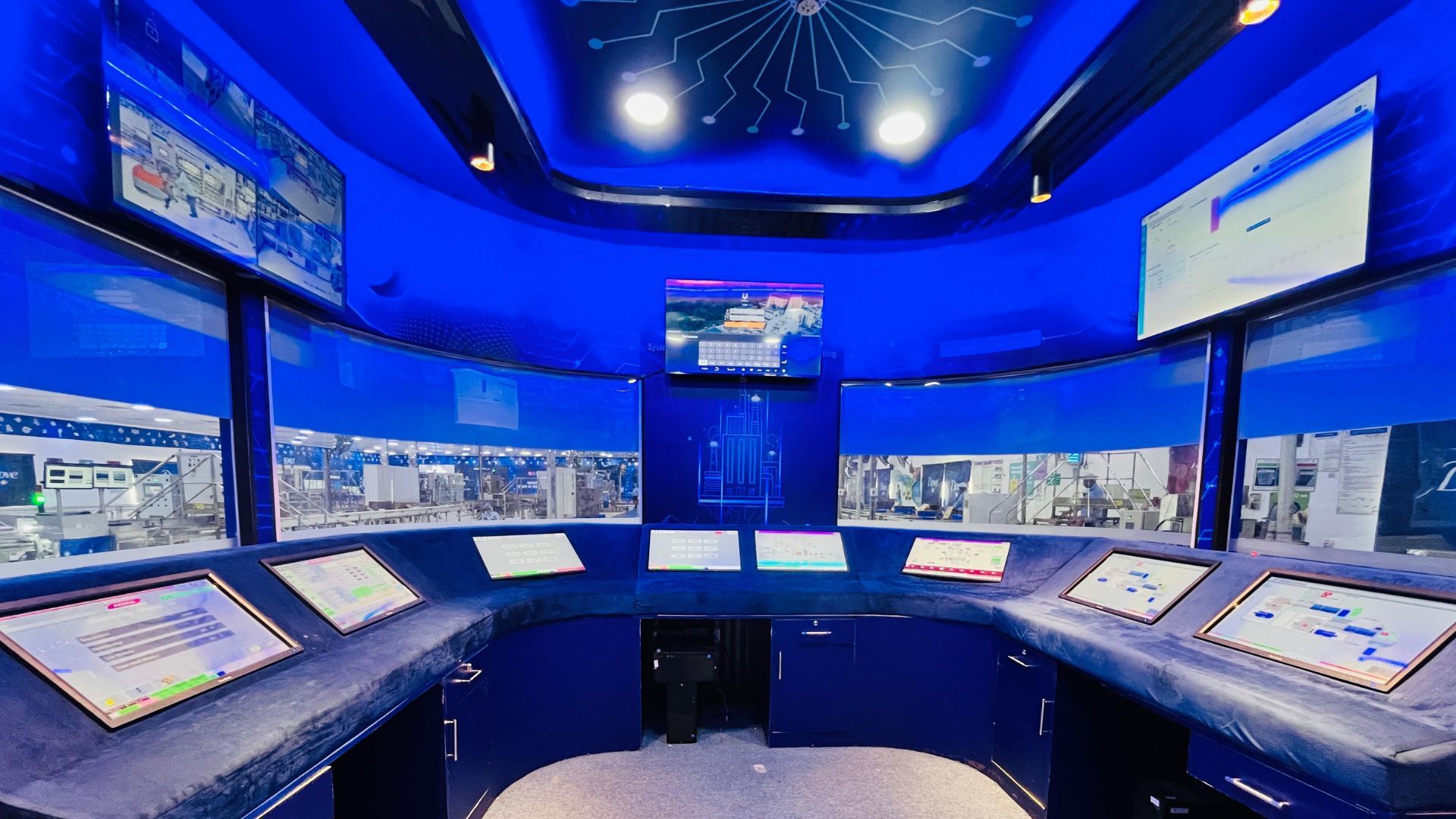 Modern control room with multiple touchscreens and monitors, set in a futuristic blue environment with a view of an automated factory floor in the background.
