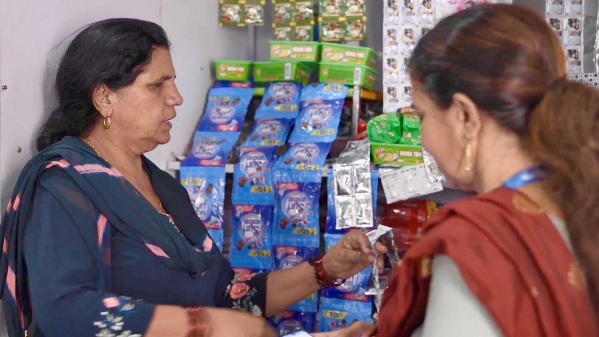Two women talking at a shop counter with various products displayed.