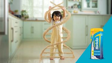 A child in a kitchen is surrounded by a DNA-shaped swirl of grains, symbolizing growth, with a Junior Horlicks container on the right.