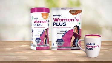 Two packs of Horlicks Women’s Plus packs in caramel and chocolate flavours featuring a woman with a spine illustration, along with a filled mug displayed on a wooden surface.