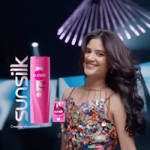 A woman with long, voluminous hair in a colorful, sequined dress stands next to a Sunsilk Thick & Long shampoo bottle and logo.