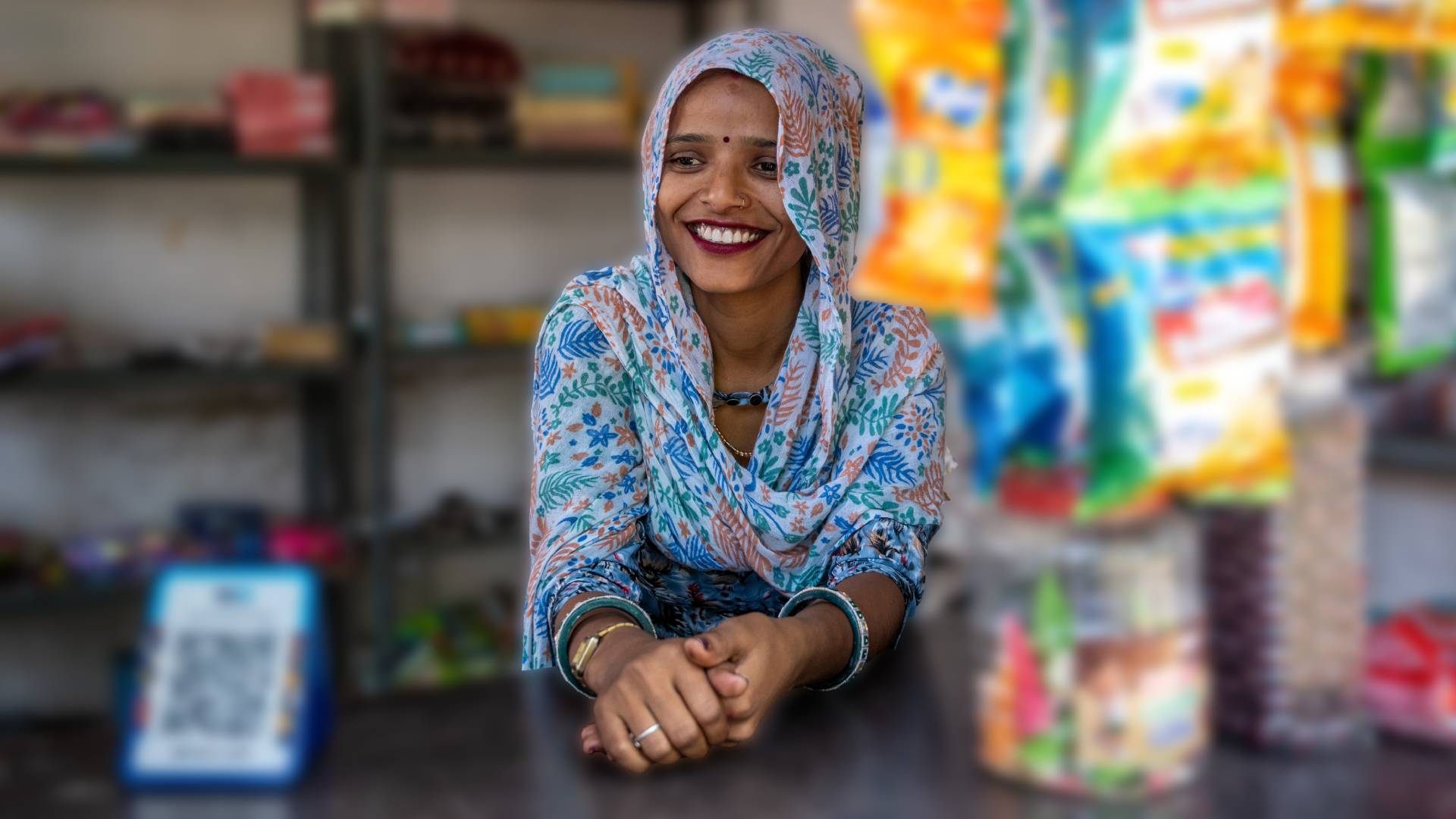 Smiling woman in floral attire at a small shop counter.